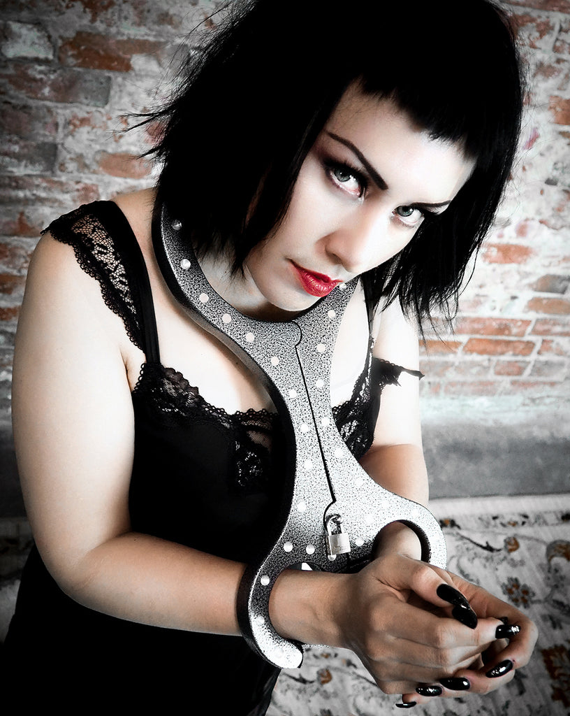 A black-haired woman in bold makeup wears a lace dress, restrained by The Cuffstore Heavy Aluminum Cangue with Padlock Bondage Yoke Shrews Fiddle Restraint, standing against a textured brick wall and gazing up at the camera.