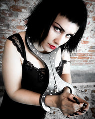 A black-haired woman in bold makeup wears a lace dress, restrained by The Cuffstore Heavy Aluminum Cangue with Padlock Bondage Yoke Shrews Fiddle Restraint, standing against a textured brick wall and gazing up at the camera.