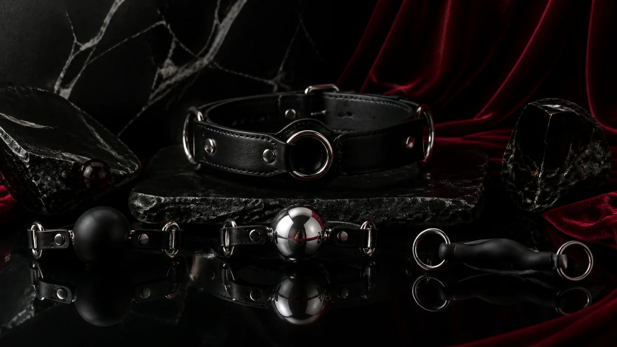 Leather harness, black silicone ball, and polished stainless gag forms on obsidian.