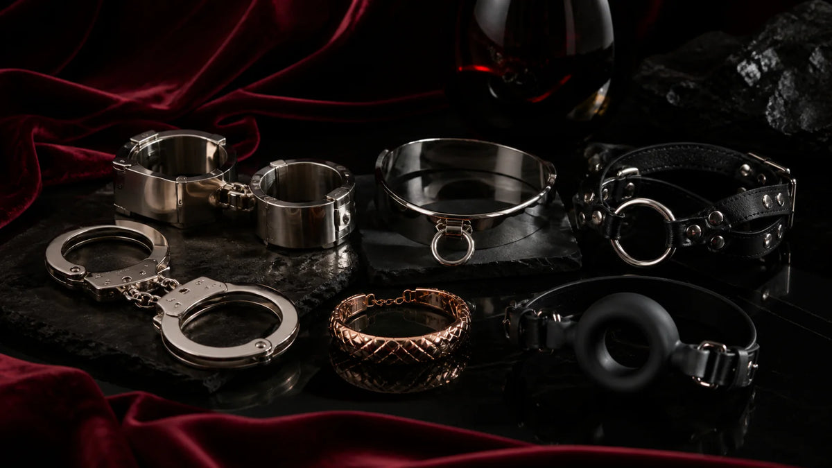 Stainless cuffs, a curved collar, rose-gold bracelet, and black gag forms on velvet.