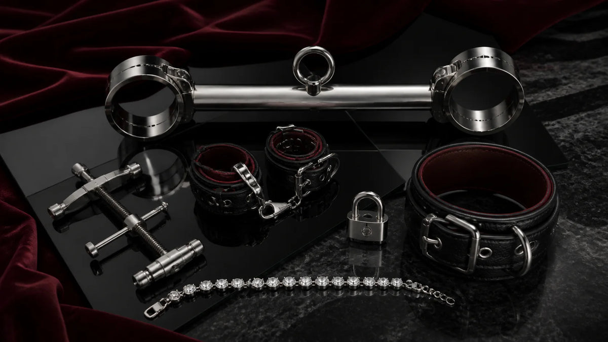 A rigid stainless spreader bar, leather restraint pieces, and a small padlock on dark stone.