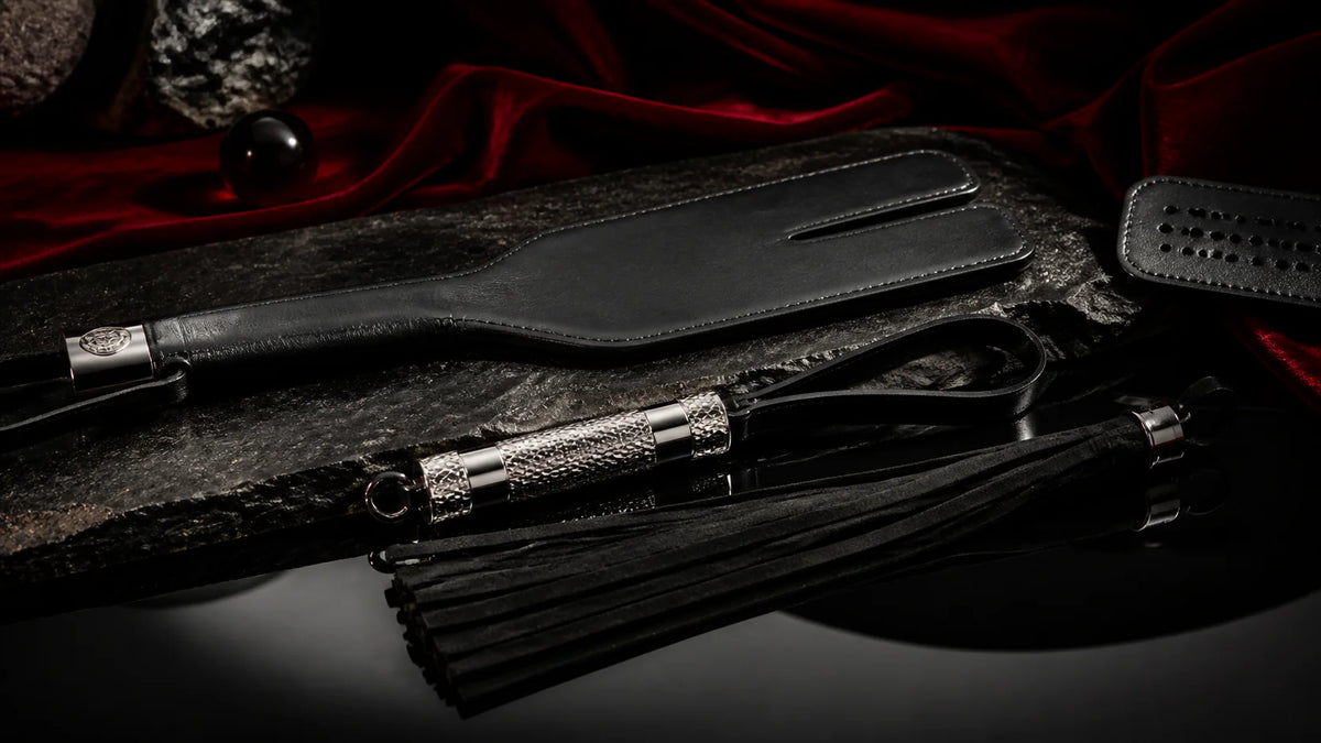 Leather paddles and a suede flogger arranged on dark stone and velvet.