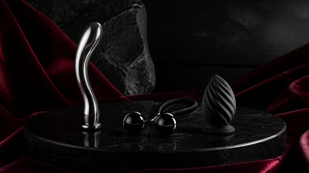 A curved stainless wand, dark silicone form, and polished clamp hardware on stone.